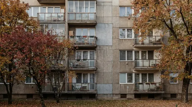 Többszintes panelház homlokzata erkélyekkel, fák között, őszi napfényben